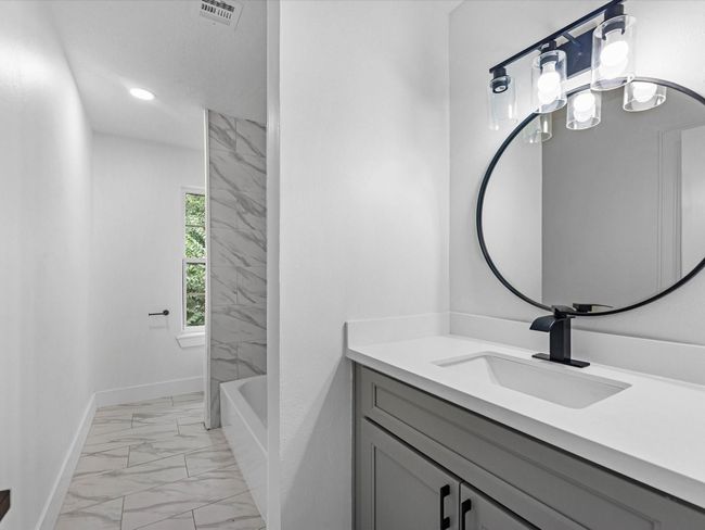Modern bathroom with a sleek vanity featuring a round mirror and stylish lighting. The space includes a tub with elegant tilework and a window for natural light. | Image 31