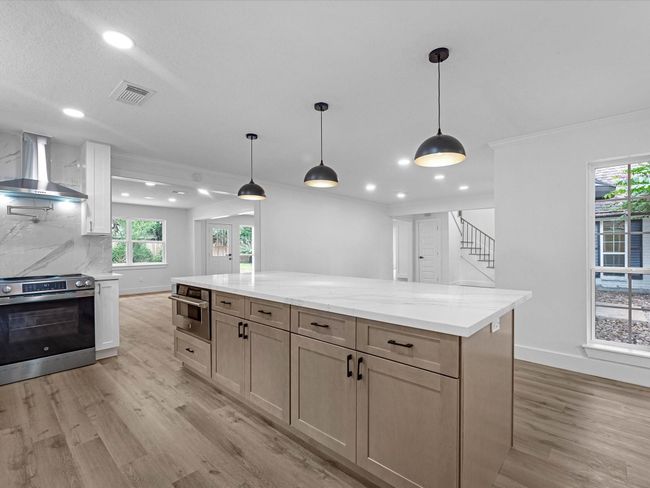 Modern kitchen with an open layout, featuring a spacious island with pendant lighting. It has wood flooring, sleek cabinetry, and stainless steel appliances. Large windows provide natural light, and there's a view of the adjacent living area. | Image 14