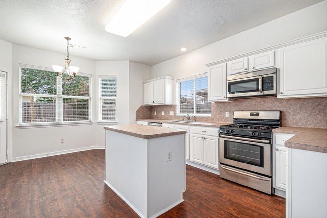 Bright kitchen with white cabinetry, stainless steel appliances, and a central island. Features wood flooring, ample natural light from large windows, and a cozy dining area with a chandelier. | Image 6