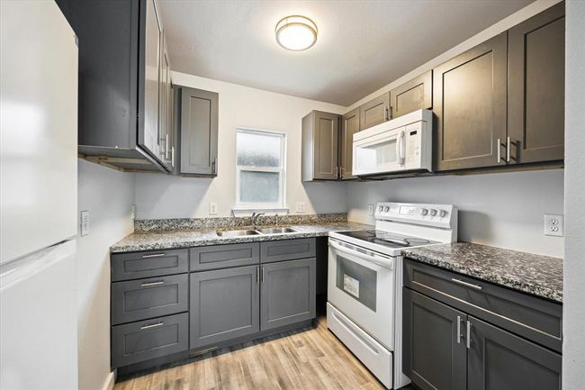 This kitchen features modern dark cabinetry, granite countertops, and a compact layout. It includes a white stove and microwave, with a window providing natural light. The space is accented by a light wood floor. | Image 5