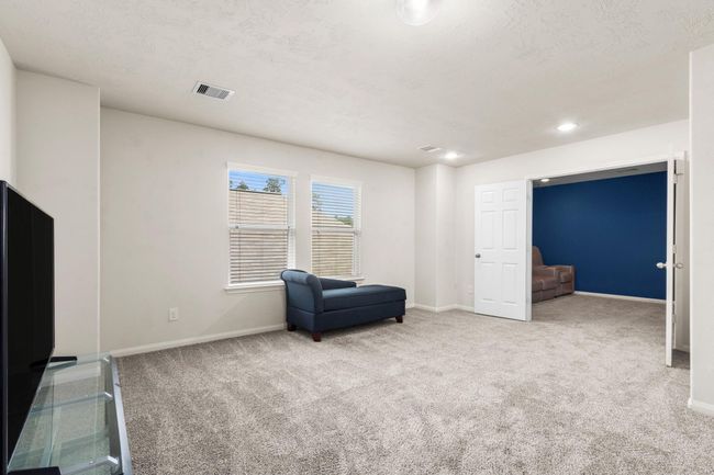 This photo showcases a spacious, carpeted living area with neutral walls, featuring a large window for natural light. It includes a cozy blue chaise and connects to an adjacent room through a doorway, offering a versatile layout. | Image 19