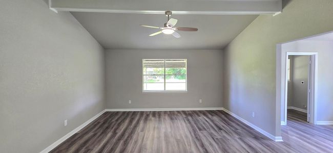 Spacious living room with vaulted ceiling, modern wood flooring, neutral gray walls, and a ceiling fan. A large window provides natural light, and an open doorway leads to adjacent rooms. | Image 12