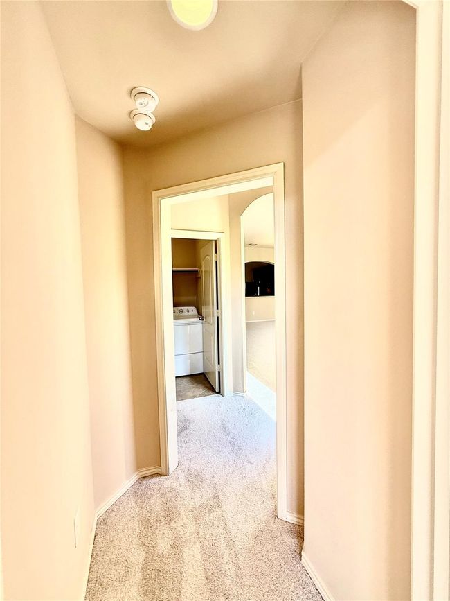When walking from main bedroom there's narrow hallway with beige walls and carpeted flooring, leading to a laundry area with a washer and dryer. Off to the right this hallway leads to the living room area. | Image 19