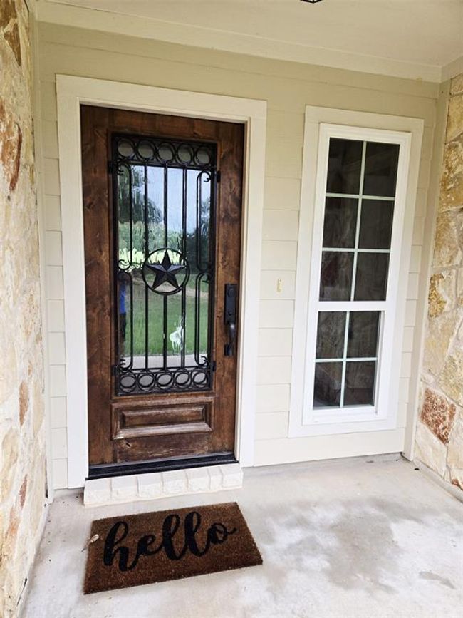 Charming entryway featuring a rustic wooden door with decorative wrought iron accents and a star motif. The adjacent tall window allows natural light. A welcoming "hello" mat sits on the concrete porch, complemented by stone and siding exterior. | Image 6