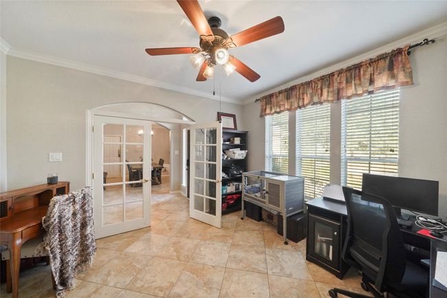 This photo showcases a bright home office with large windows, tiled flooring, and a ceiling fan. The room features French doors leading to an adjacent space, enhancing its openness and connectivity. Ideal for a productive work environment. | Image 8