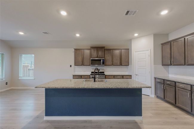 Kitchen featuring decorative backsplash, visible vents, stainless steel appliances, light stone countertops, and an island with sink | Image 18