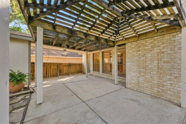 This photo showcases a spacious, paved patio area with a wooden pergola, offering partial shade. The space is adjacent to the house with large windows, providing a great spot for outdoor relaxation or entertaining. | Image 22