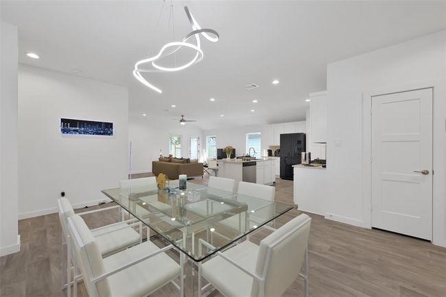 This photo showcases a modern, open-concept living space with a sleek glass dining table and contemporary lighting. The layout flows seamlessly from the dining area to the kitchen and living room, featuring white cabinetry, a black refrigerator, and wood flooring, creating a bright and inviting atmosphere. | Image 6