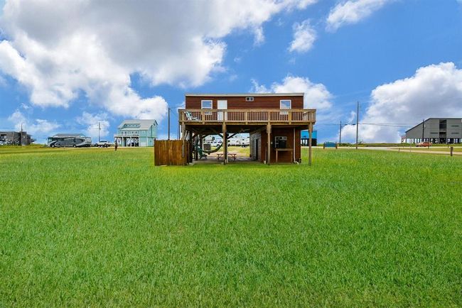 This photo shows a stilted wooden house with a spacious deck, set on a large grassy lot. The area is open with a few neighboring homes, under a bright blue sky with scattered clouds. Ideal for those seeking a serene, beach setting. | Image 21