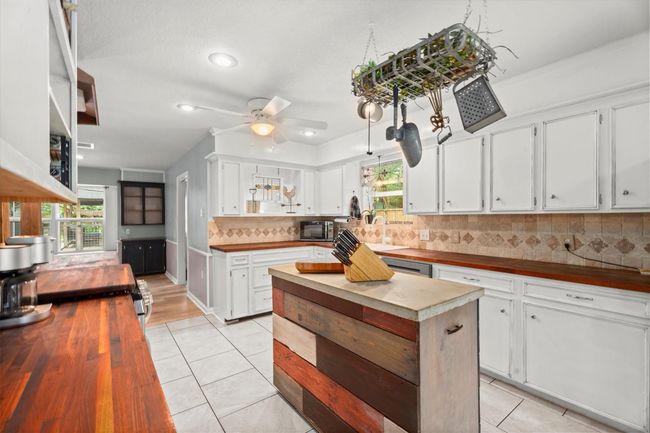 This kitchen features white cabinetry, a stylish tile backsplash, and a central island with a rustic wood finish. Wooden countertops add warmth, while a hanging pot rack enhances functionality. Large windows provide ample natural light, creating an inviting space. | Image 10