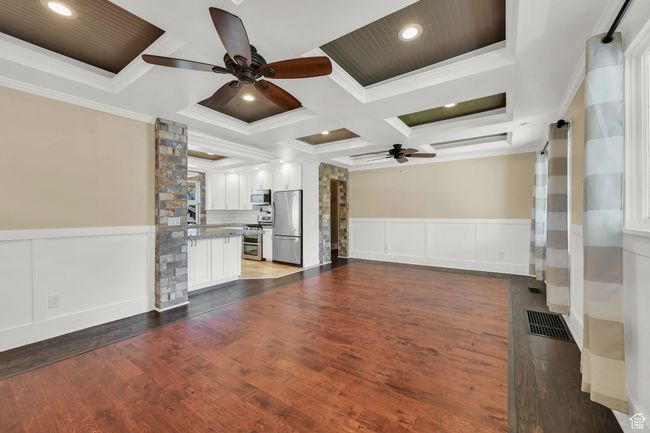Unfurnished living room with a ceiling fan, a wainscoted wall, coffered ceiling, ornamental molding, and visible vents | Image 6
