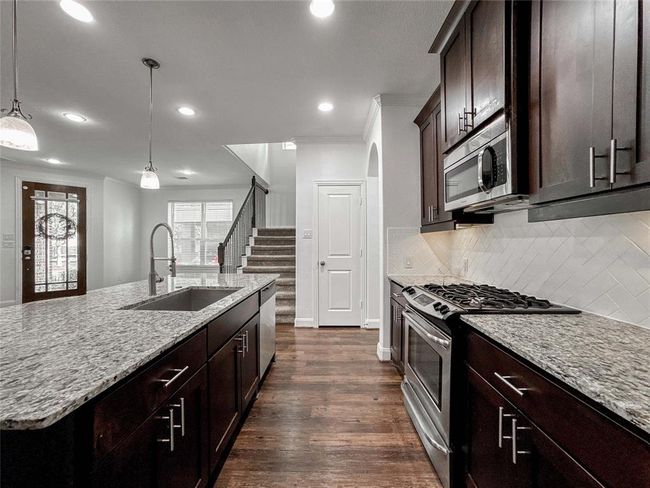 This modern kitchen features sleek dark wood cabinets, granite countertops, and stainless steel appliances. A large island with a sink provides ample prep space. The open layout leads to a bright living area with a staircase, enhancing the home's contemporary appeal. | Image 11