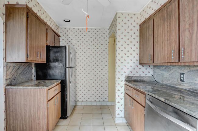 Kitchen featuring brown cabinetry, wallpapered walls, light tile patterned flooring, decorative backsplash, and appliances with stainless steel finishes | Image 11