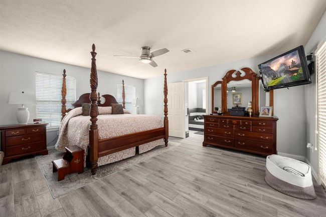 This is a spacious bedroom featuring a large four-poster bed, matching wooden furniture, and modern grey flooring. The room includes a ceiling fan, ample natural light from two windows, and is connected to an ensuite bathroom. A mounted TV provides in-room entertainment. | Image 36