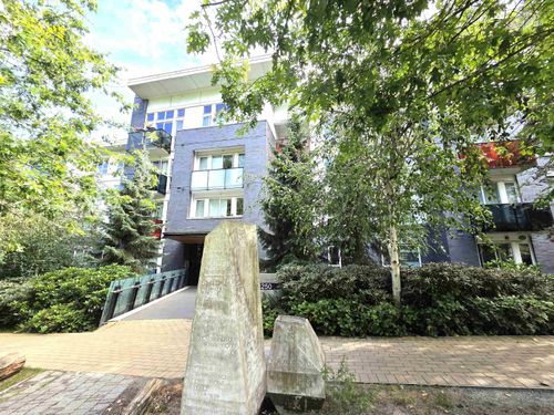Multi-story residential building featuring dark brick cladding, multiple balconies with glass railings, and a prominent entry bridge with glass panels | Card Image