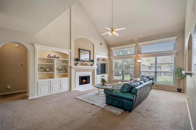 The family room boasts soaring two-story ceilings, creating a spacious and airy atmosphere. A gas-started fireplace is framed by built-ins on both sides, while large windows along the back wall, topped with transom windows, flood the room with natural light. An arched doorway to the left leads to the primary suite, and to the right, you can see the overhang of the breakfast bar that connects to the kitchen. | Image 9