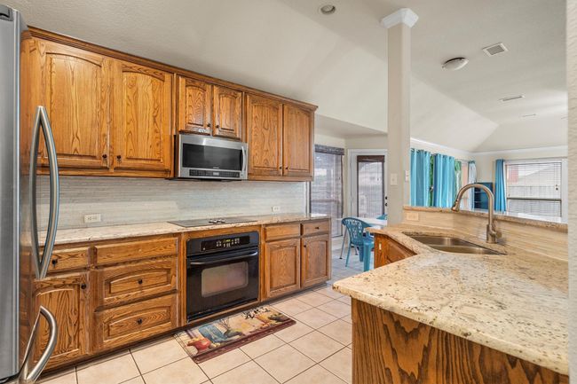 Kitchen featuring black appliances, decorative backsplash, brown cabinetry, light stone countertops, and lofted ceiling | Image 16