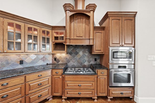 Kitchen featuring stainless steel appliances, tasteful backsplash, glass insert cabinets, dark stone counters, and brown cabinets | Image 11
