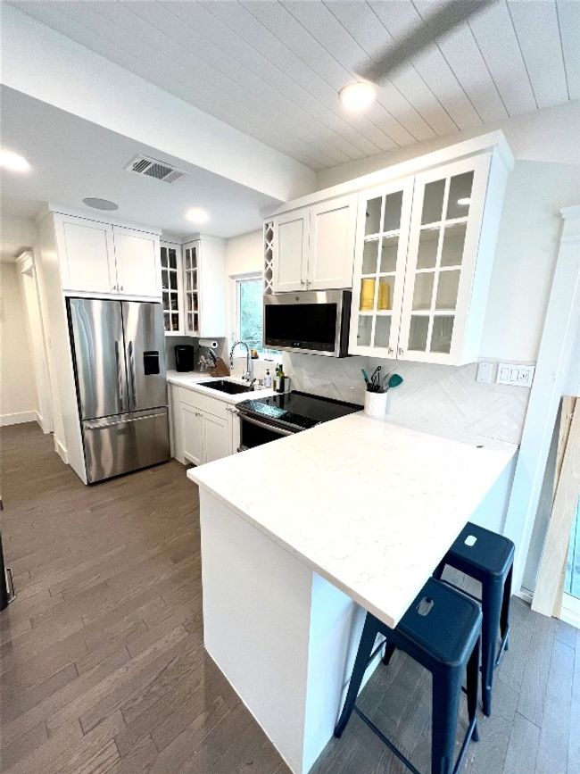 This kitchen features a modern design with sleek stainless steel appliances, white cabinetry with glass doors, and a quartz countertop. The space is bright and functional, with a cozy breakfast bar and a clean, contemporary look. | Image 6