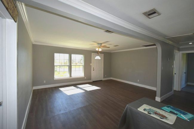 This spacious living area features dark wood floors, gray walls, and abundant natural light from large windows. It connects to an open kitchen with modern appliances, creating a welcoming and versatile space. | Image 6