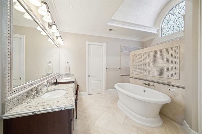 This elegant bathroom features a freestanding tub, marble countertops, and a large mirror. The space is enhanced by natural light from a decorative arched window, creating a luxurious and serene atmosphere. | Image 23