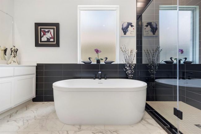 This contemporary bathroom features a sleek freestanding tub, elegant black tile accents, and a stylish vanity. The space is complemented by artistic wall decor and a frosted window for privacy and natural light. | Image 30