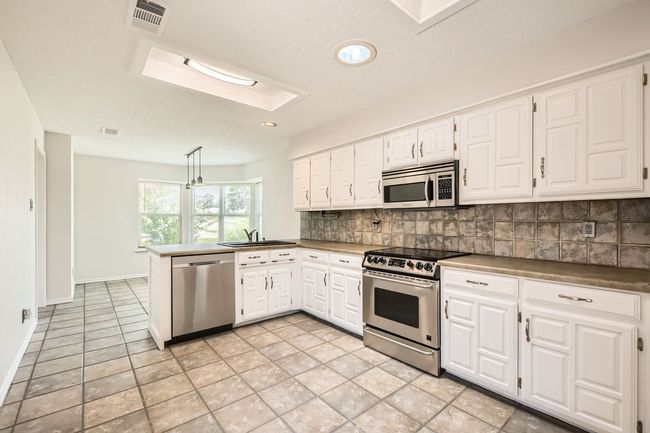 Kitchen with appliances with stainless steel finishes, a peninsula, a skylight, decorative backsplash, and white cabinetry | Image 7