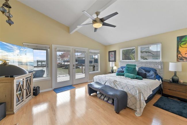 Bright and airy primary bedroom with vaulted ceilings, featuring large windows and glass doors opening to a waterfront view. Includes a ceiling fan, hardwood floors, and modern furnishings, creating a relaxing and stylish retreat. | Image 27