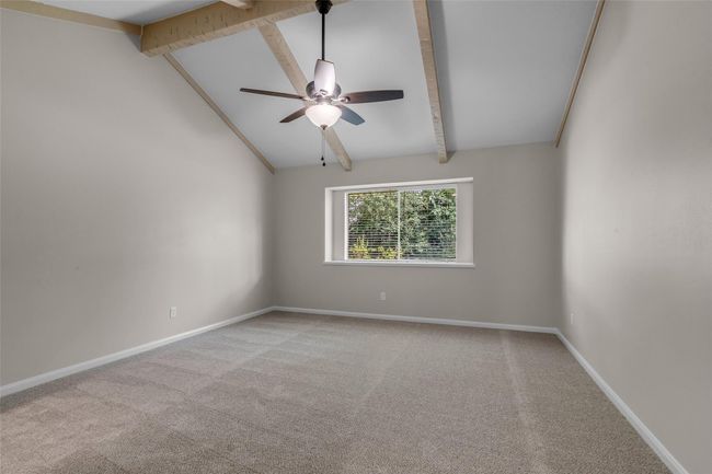 This spacious primary bedroom features a vaulted ceiling with exposed wooden beams, a ceiling fan, neutral-colored walls, and a large window offering plenty of natural light. Carpeted floors add warmth and comfort. | Image 25