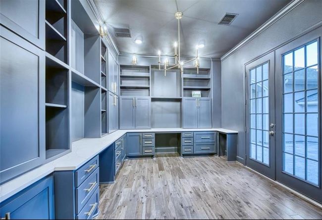 This photo showcases a sophisticated home office with built-in blue cabinetry, ample shelving, and a stylish chandelier. The space is brightened by large glass doors, and it features elegant wood flooring, providing a modern and functional work environment. | Image 15