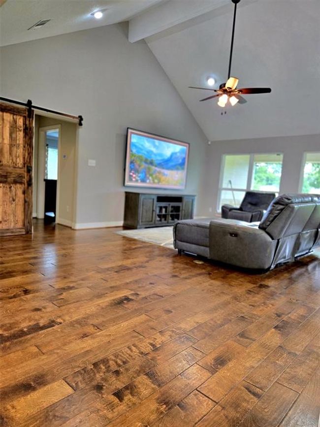 Spacious living room with high vaulted ceilings, hardwood floors, and a ceiling fan. Features a large TV, cozy recliner seating, and natural light from multiple windows. Includes a rustic sliding barn door for a stylish touch. | Image 16