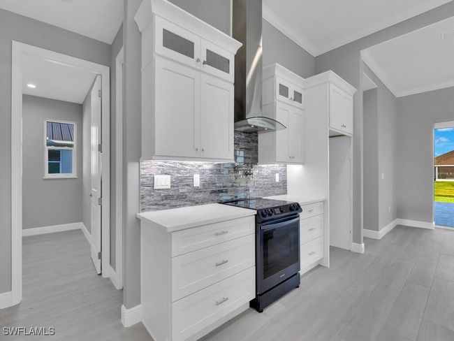 Kitchen featuring electric range, wall chimney range hood, decorative backsplash, glass insert cabinets, and light countertops | Image 12