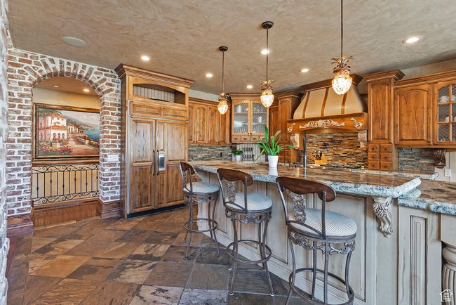 Kitchen with brown cabinetry, custom range hood, glass insert cabinets, and stone tile flooring | Image 22