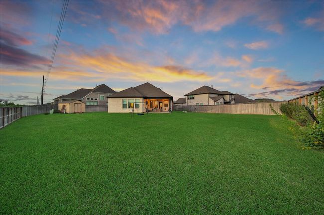 Expansive backyard with lush grass, bordered by a tall wooden fence for privacy. The single-story house features a covered patio, perfect for outdoor relaxation. The sky is beautifully lit with a colorful sunset, enhancing the serene atmosphere. | Image 10