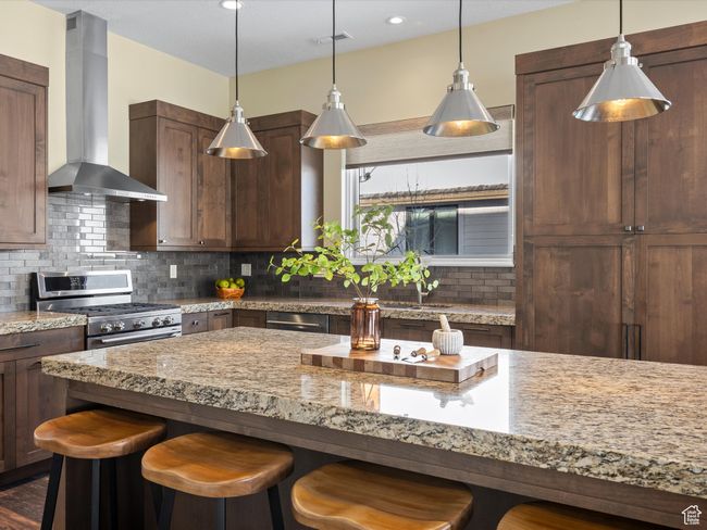 Kitchen featuring wall chimney range hood, stainless steel range with gas stovetop, a breakfast bar, tasteful backsplash, and light stone counters | Image 8