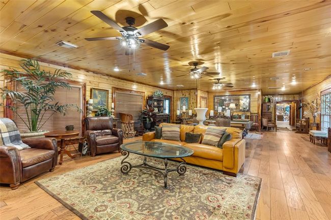 This spacious living room features warm wood paneling, a cozy seating area with leather chairs and a sofa, ceiling fans, and plenty of natural light. The open layout is perfect for entertaining, with elegant furnishings and a rustic charm. | Image 37
