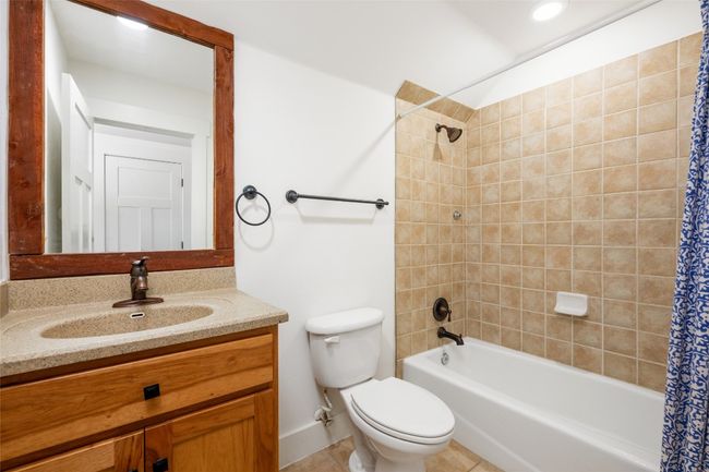 The guest bath features a tiled shower and bathtub, tile floor, natural wood vanity with solid surface counter, and a framed mirror. | Image 31