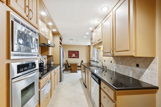 Galley-style kitchen featuring modern stainless steel appliances, ample cabinetry, and granite countertops. A serving hatch connects it to the adjacent room, enhancing the open feel. | Image 12
