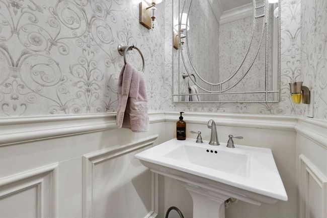Designed with timeless appeal, the downstairs half bath features tasteful wallpaper above crisp white wainscoting and a traditional chair rail, creating a refined and welcoming ambiance. A pedestal sink adds graceful simplicity, while decorative lighting and a stylish framed mirror complete the look—adding a touch of true elegance. | Image 39