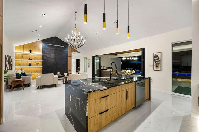 The kitchen features a bold waterfall island with luxurious black granite countertops and natural wood cabinetry, accented by contemporary pendant lighting. Large sliding glass doors seamlessly connect the indoor space to an inviting outdoor area, perfect for entertaining. Polished marble flooring and integrated lighting throughout enhance the bright, airy atmosphere, while a striking feature wall adds a sophisticated focal point to the living space. | Image 9