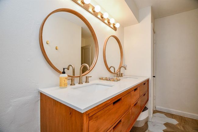This bathroom features a modern double vanity with sleek wooden cabinetry and white countertops. It includes two round mirrors and contemporary faucets, offering a stylish and functional space. Ample lighting is provided by a row of bulbs above the mirrors. | Image 38