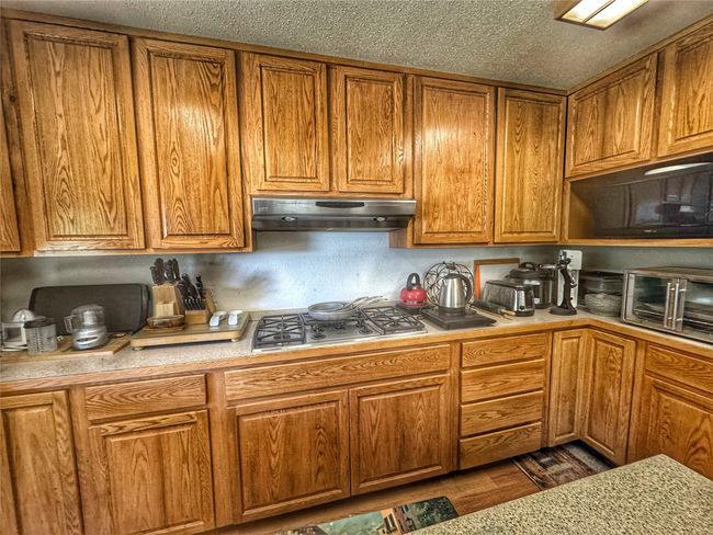 Kitchen featuring brown cabinetry, a textured ceiling, stainless steel gas cooktop, under cabinet range hood, and light countertops | Image 14