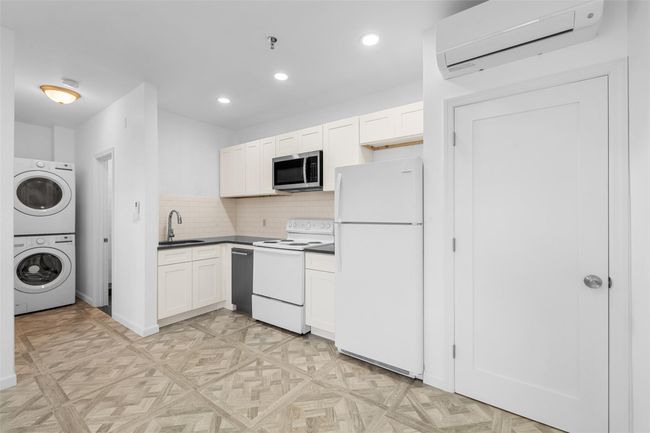 Kitchen featuring a wall unit AC, stacked washer and dryer, white appliances, a sink, and dark countertops. | Image 5