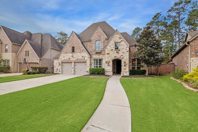 This is an elegant two-story brick and stone home featuring a well-maintained lawn, a two-car garage, and a welcoming arched entryway, nestled in a wooded residential neighborhood with mature trees. | Image 2