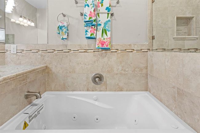 This is a modern bathroom featuring a large built-in bathtub with whirlpool jets, surrounded by neutral-toned tile. Above the tub is a well-lit vanity mirror, and the space is accented with a colorful towel, adding a touch of vibrancy. | Image 31