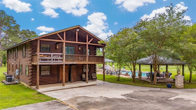 This photo showcases a charming two-story log cabin with a spacious balcony, set in a lush, green landscape. To the right, there's a pool area with a covered patio, perfect for outdoor entertaining. The setting is serene and inviting, ideal for those seeking a rustic yet comfortable home. | Image 44