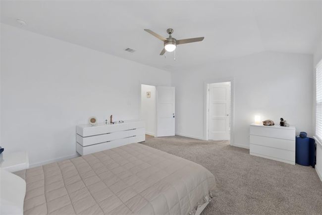 This spacious bedroom features neutral carpeting, a ceiling fan, and ample natural light. It includes modern white dressers and a large bed, creating a clean and inviting atmosphere. | Image 13