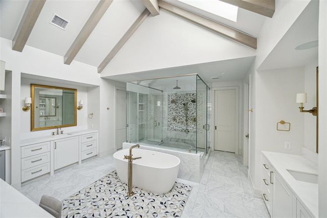 The serene primary bath is a true oasis, featuring majestic white pine ceiling beams with LED trim details that enhance its tranquil ambiance. Skylights flood the vaulted ceiling with natural light, further elevating the sense of relaxation and luxury. Custom cabinetry, separate vanities with luxe gold hardware compliment ample space for toiletries and more. | Image 17