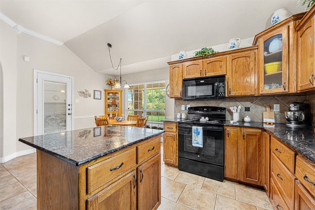 Kitchen with black appliances, vaulted ceiling, light tile patterned flooring, decorative backsplash, and brown cabinets | Image 15