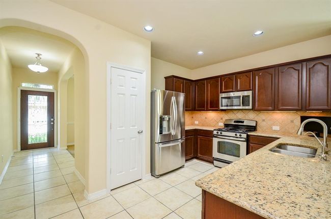 This kitchen features granite countertops, stainless steel appliances, and rich wood cabinetry. It has a tiled floor and an open layout, leading to a well-lit entrance hallway with a modern front door. | Image 11
